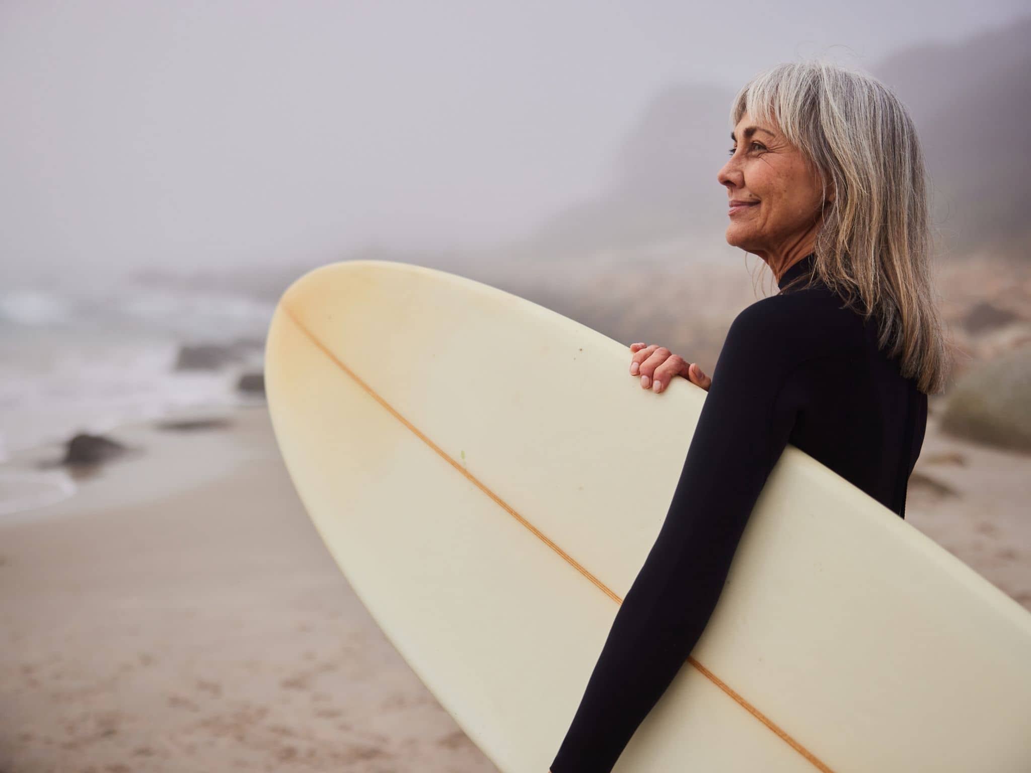 Postmenopause: Eine Frau mit grauen Haaren mit Surfbrett am Strand