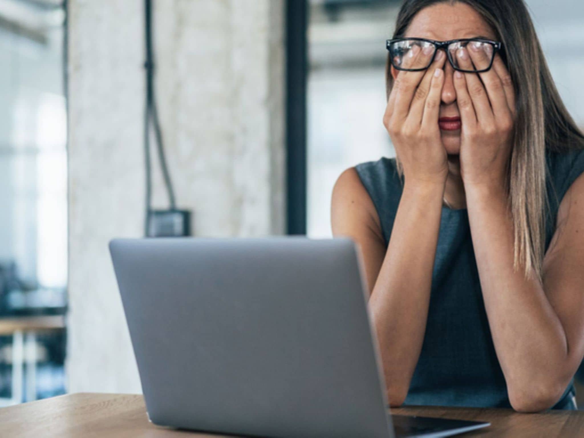 Augenringe: Frau mit Brille reibt sich die Augen, während sie am Schreibtisch vor einem Laptop sitzt