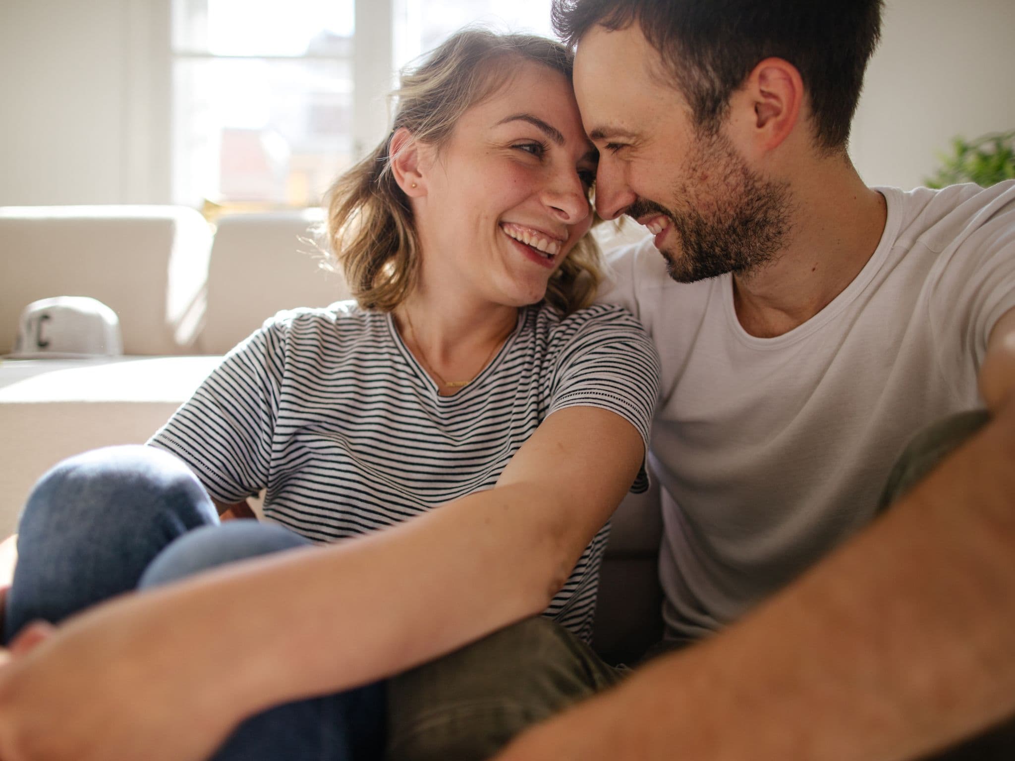 Cunnilingus: Eine junge Frau und ein junger Mann sitzen auf dem Fußboden im Schlafzimmer und sehen sich verliebt in die Augen.