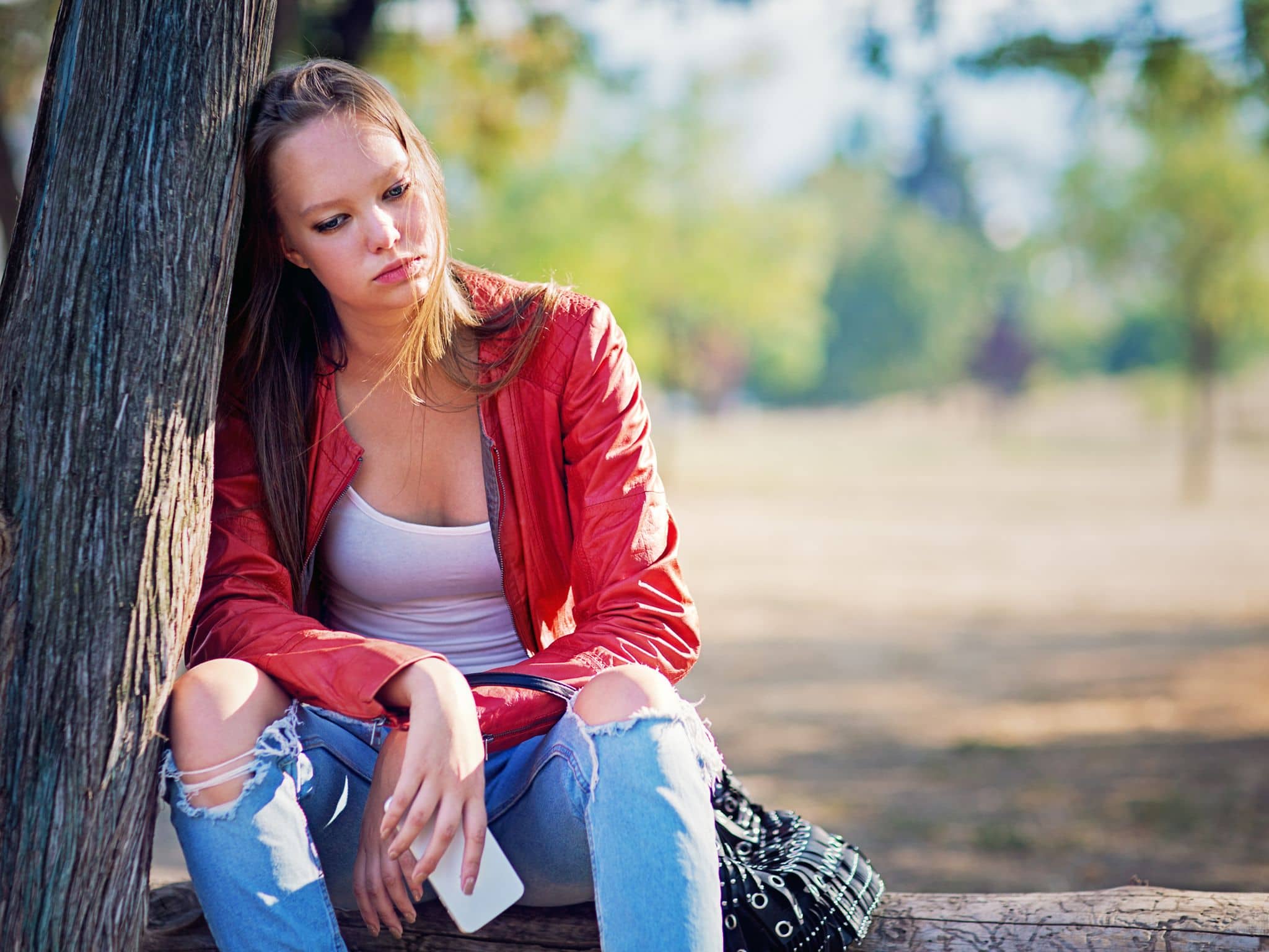 Unerwiderte Liebe: Ein junges Mädchen sitzt an einem Baum und schaut traurig ins Leere, ihr Handy hält sie in der Hand
