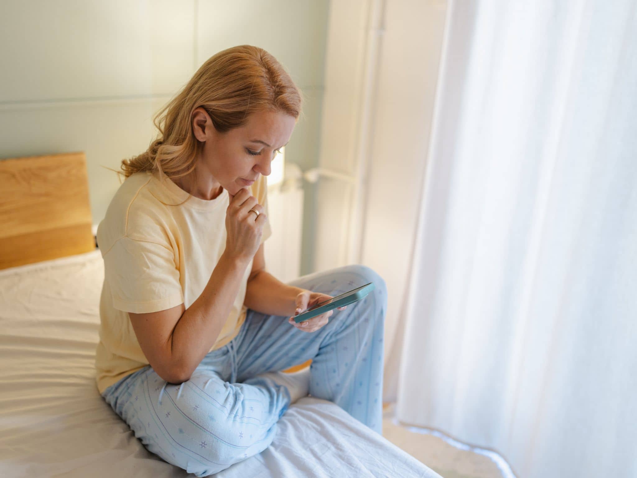 Einnistungsblutung oder Periode: Eine Frau sitzt auf einem Bett und blickt besorgt auf ihr Smatphone.