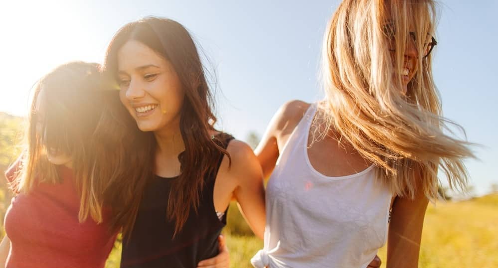 Drei junge Frauen stehen lachend eng beieinander auf einer sonnigen Wiese und umarmen sich freundschaftlich.