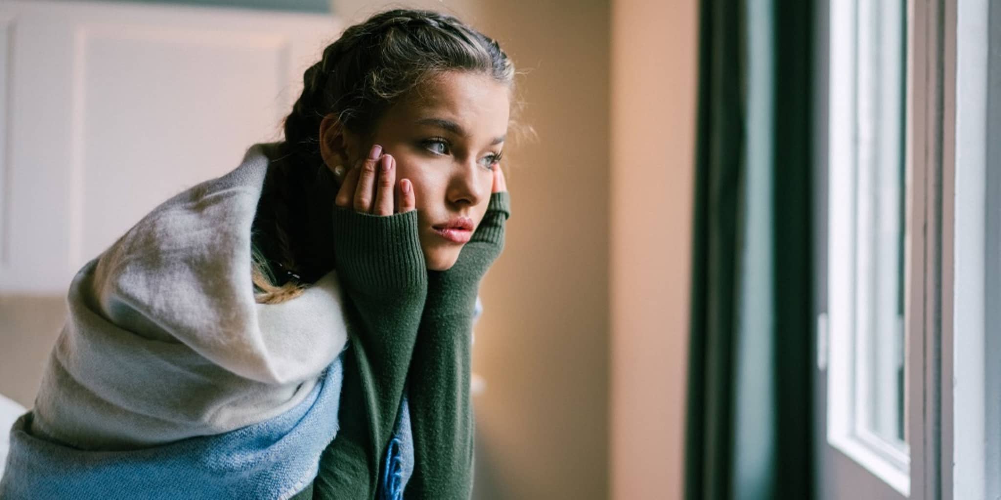 Ein Mädchen sitzt nachdenklich am Fenster, das Gesicht ist verdeckt. Sie trägt einen Schal und blickt nach draußen.