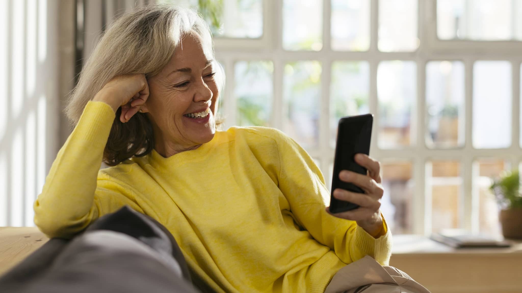 Eine ältere Frau mit gelben Pullover schaut auf Ihr Smartphone und lächelt.