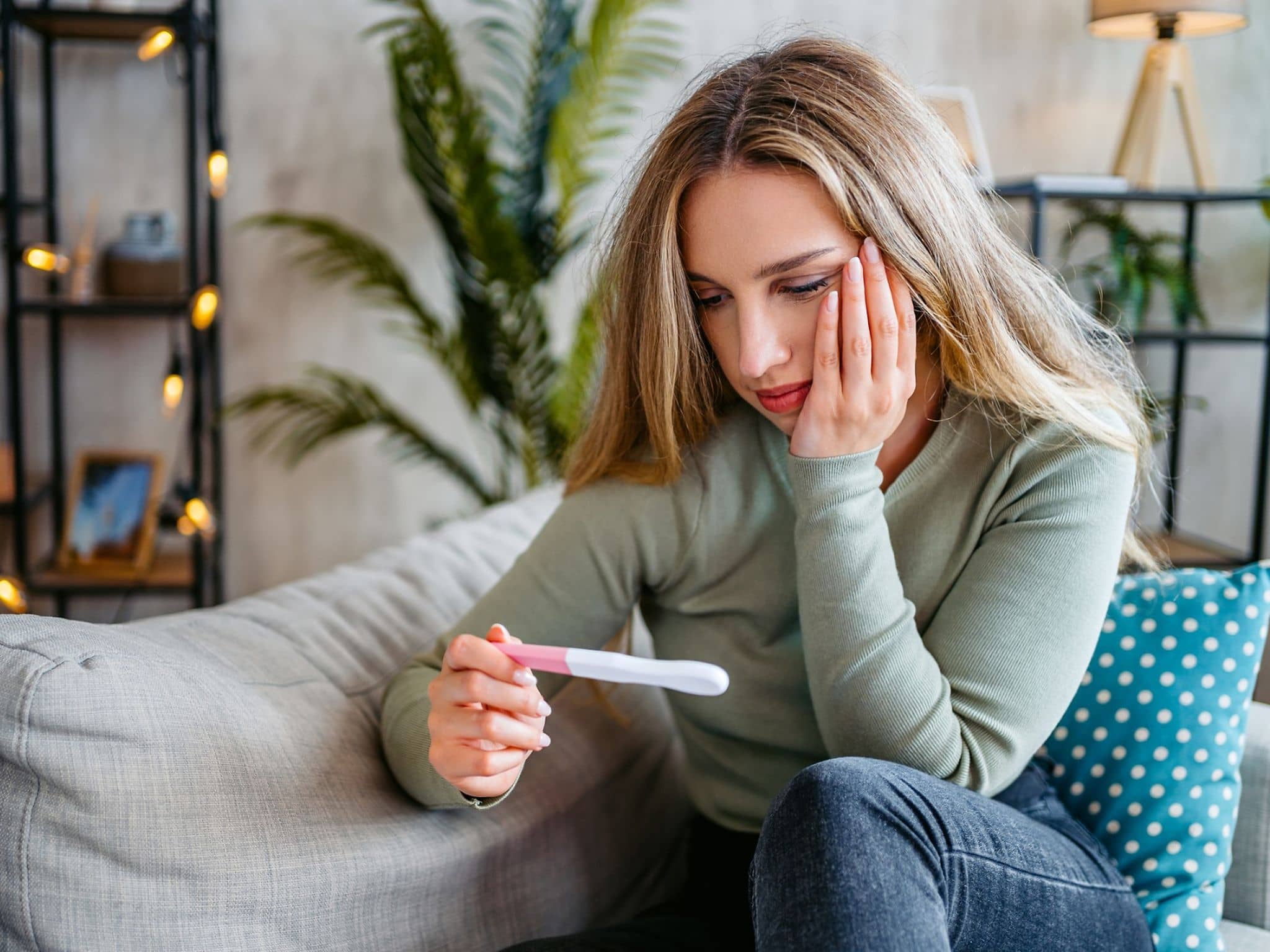 Junge Frau sitzt auf dem Sofa, hält einen Schwangerschaftstest in der Hand und schaut nachdenklich darauf.