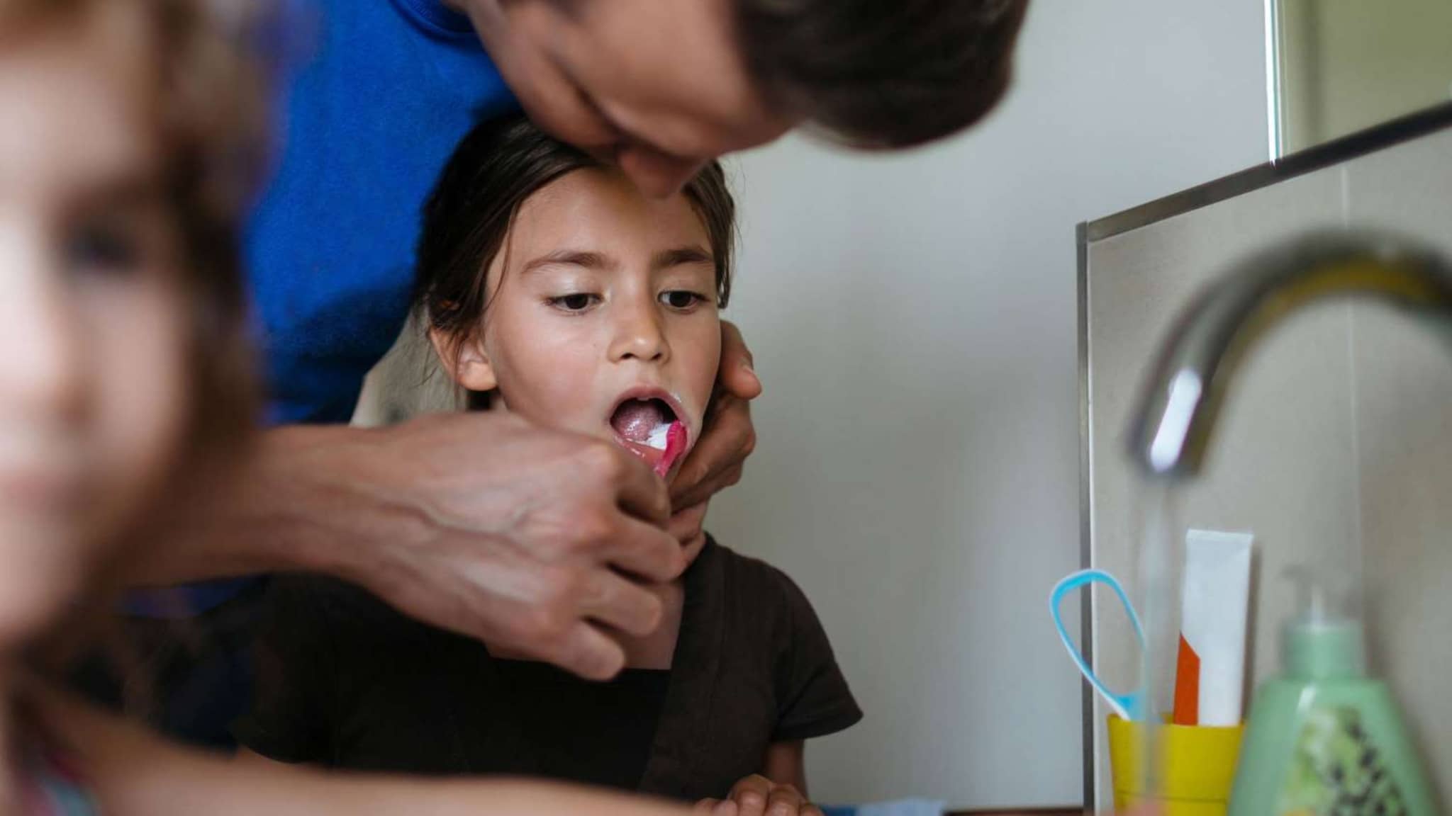 Zähneputzen: Vater putzt seiner Tochter am Waschbecken mit einer roten Zahnbürste die Zähne.