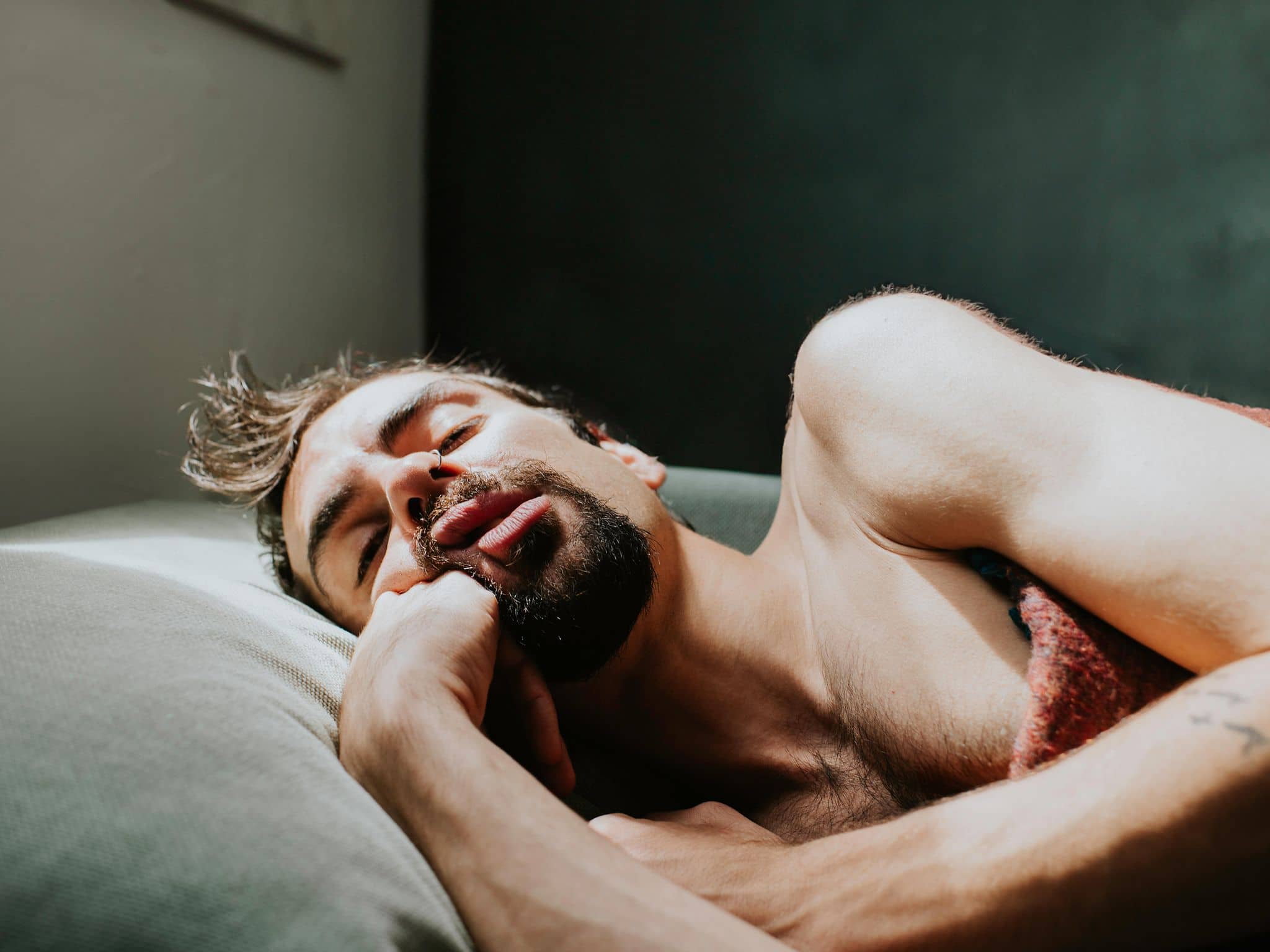 Mann schläft oberkörperfrei, halb zugedeckt und mit offenem Mund im Bett.