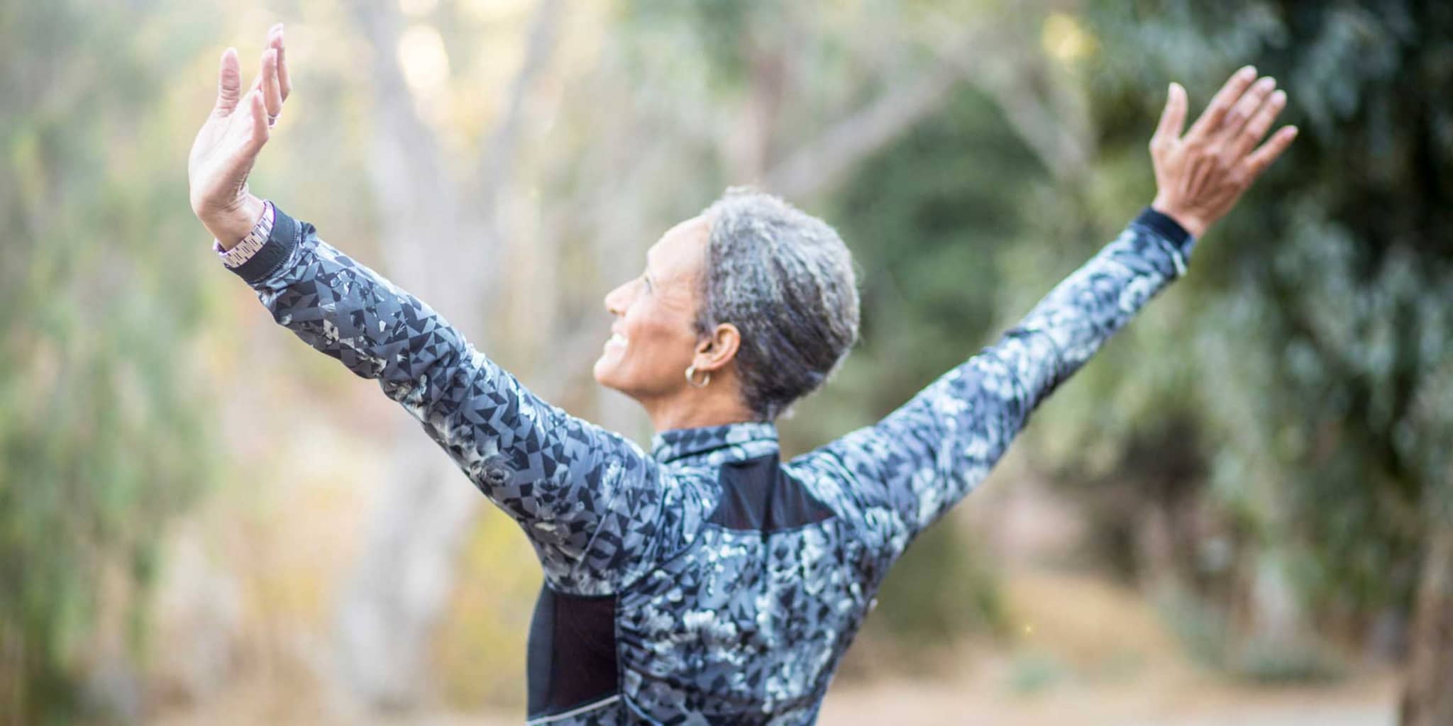 DAK-Rückentraining für Senioren: Ältere Frau in Sportkleidung streckt draußen ihre Arme seitlich aus.