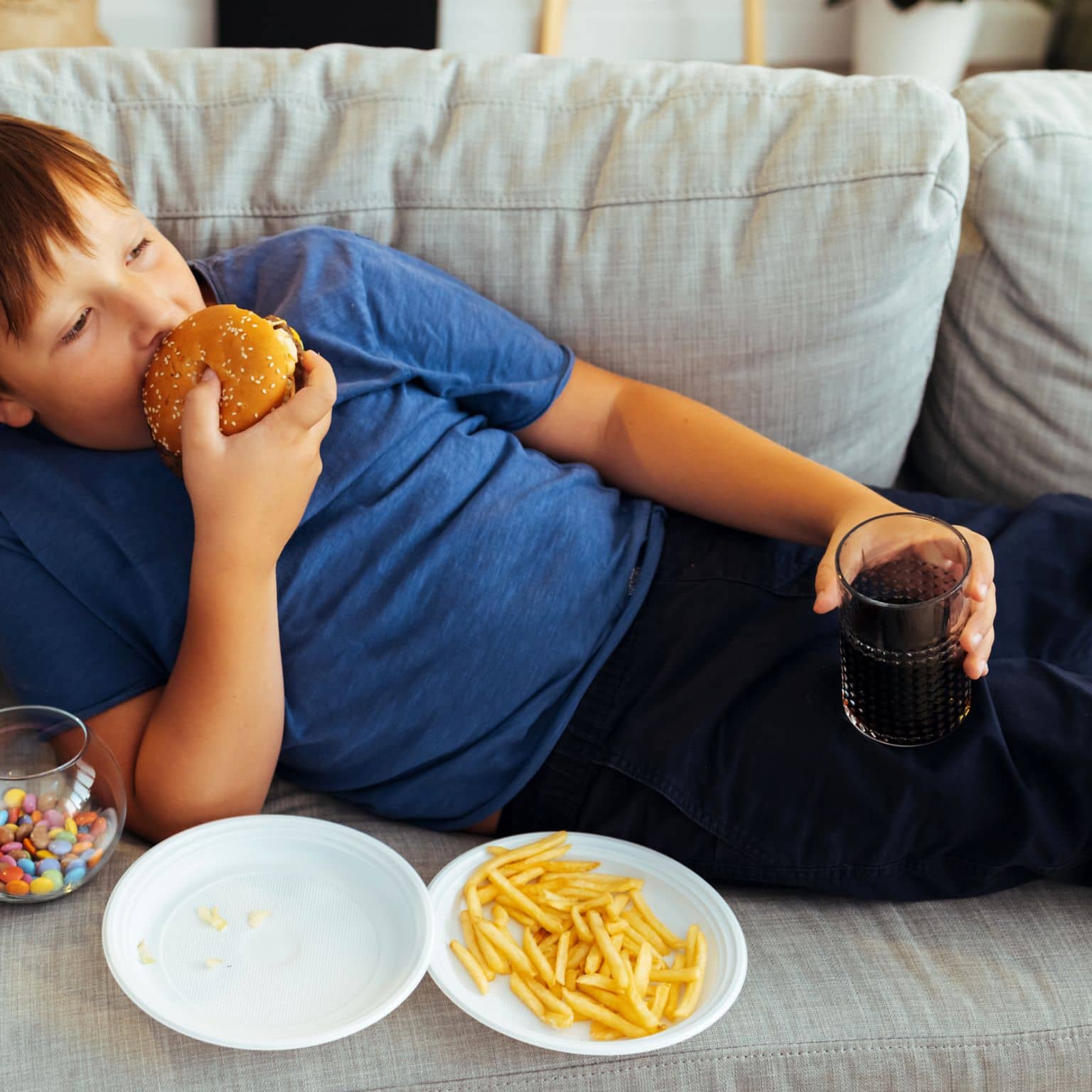 Adipositas: Ein kleiner Junge liegt auf dem Sofa und isst einen Hamburger und Pommes Frites.