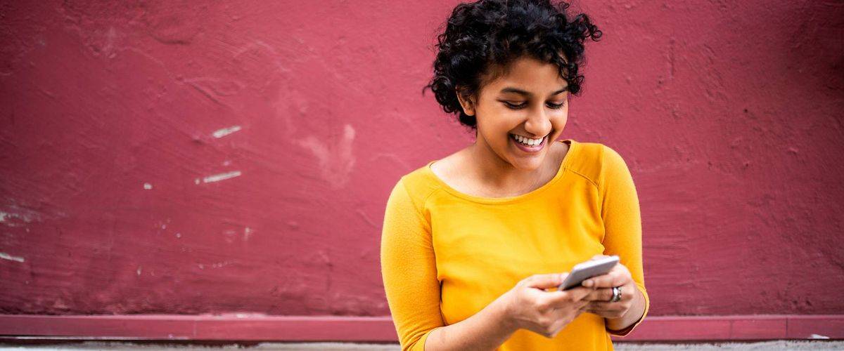 Bild: Junge Frau vor einer farbenfrohen Wand schaut auf Ihr Handy und freut sich.