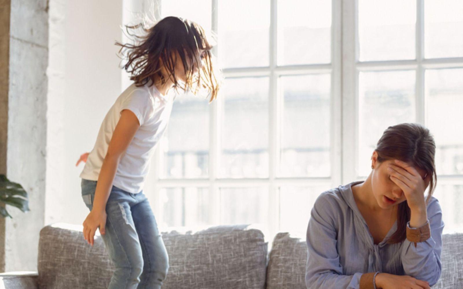 Erziehung ohne Strafen: frustrierte Mutter sitzt auf der Couch, während ihr Kind auf der Couch herum springt