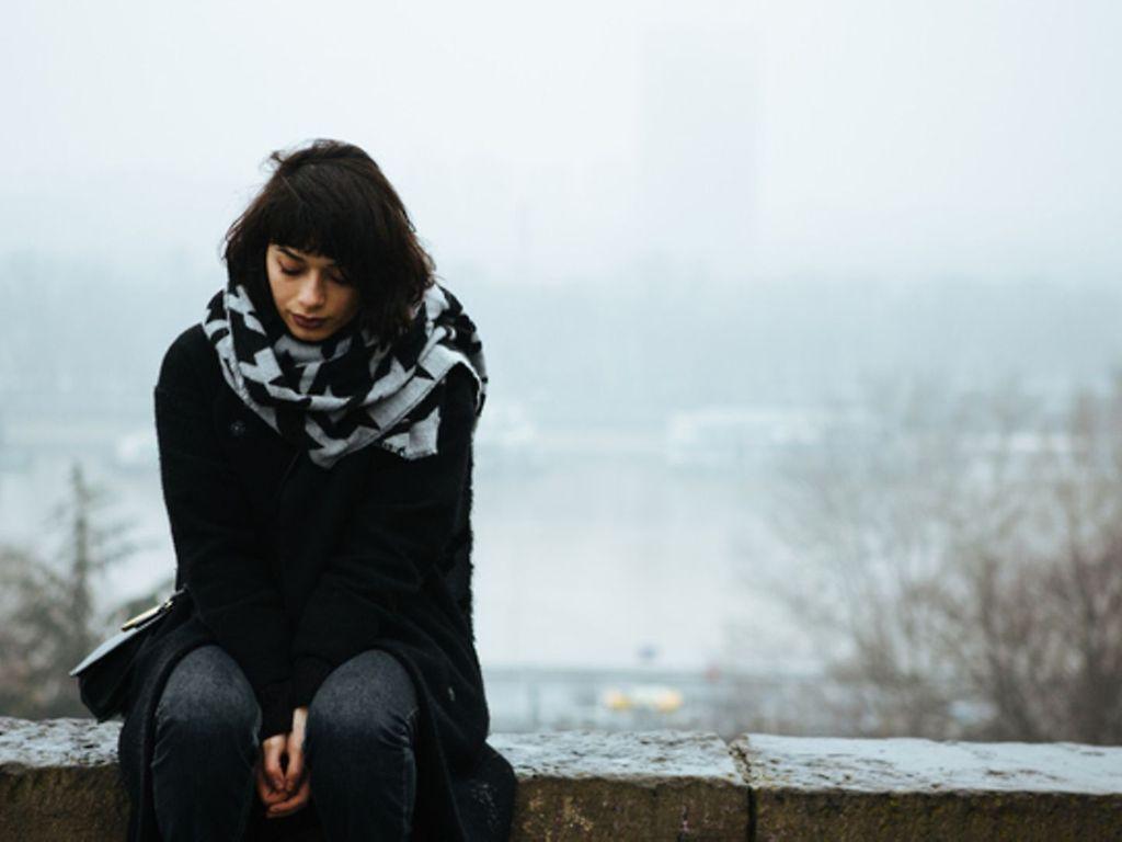 Winterdepression: Frau sitzt in einer Winterlandschaft traurig auf einer Mauer.