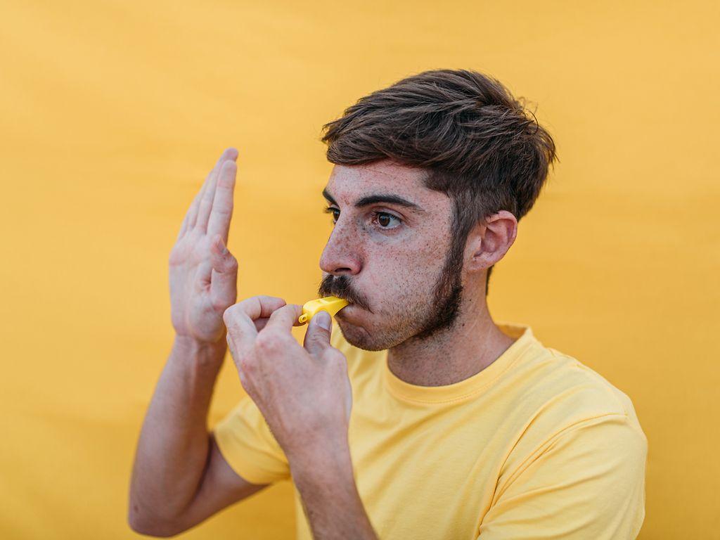 Ein junger Mann bläst in eine gelbe Pfeife, wie ein Schiedsrichter. Er ist gelb gekleidet, und auch der Hintergrund ist gelb.