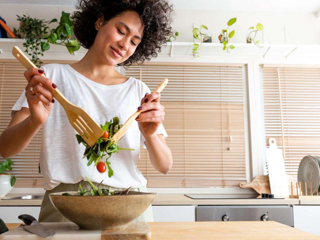 Ernährungs-Tipps: Junge Frau steht in der Küche und bereitet einen Salat zu.