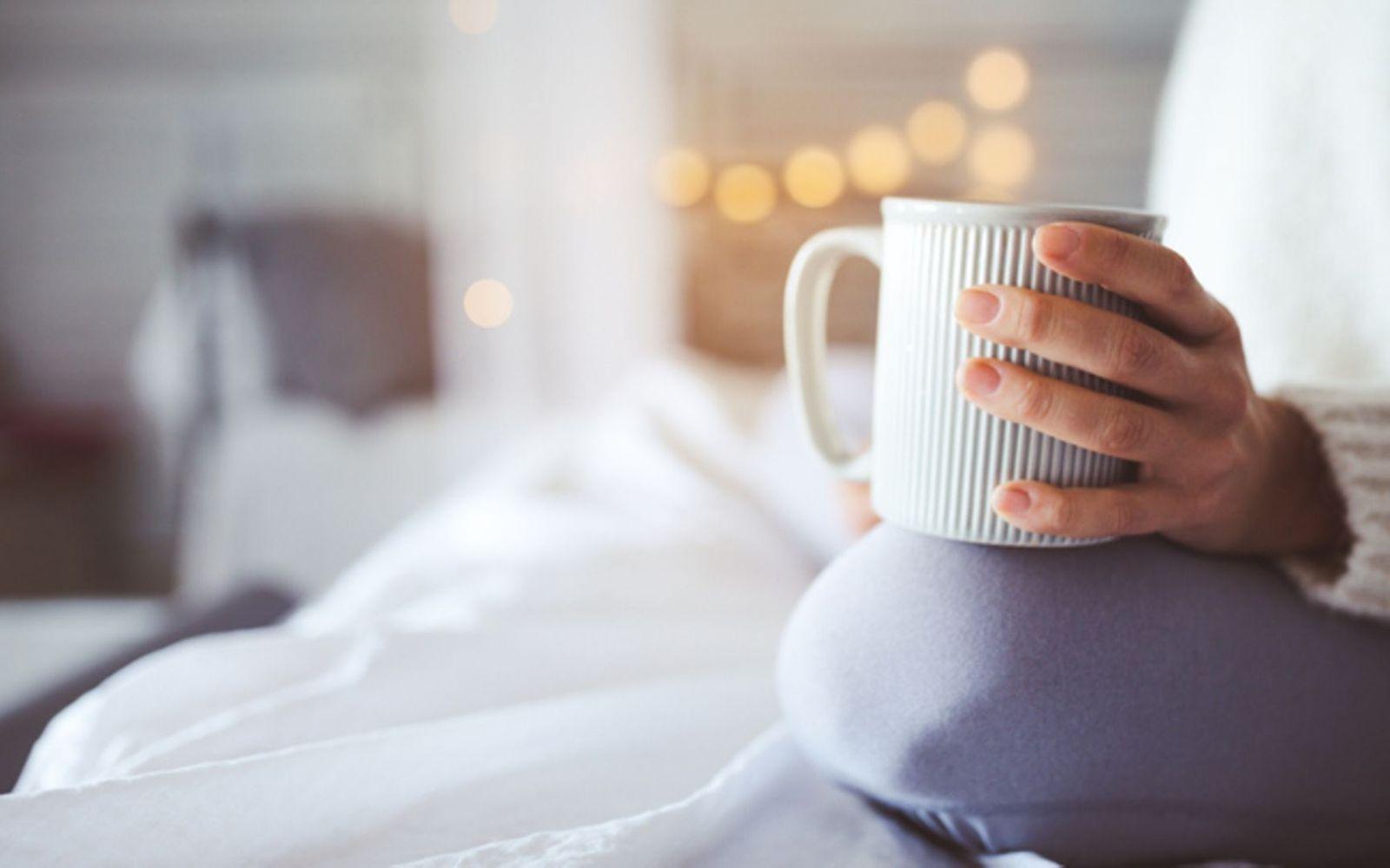 Morgenrituale: Eine Frau sitzt im Bett und hält eine Kaffeetasse in der Hand.