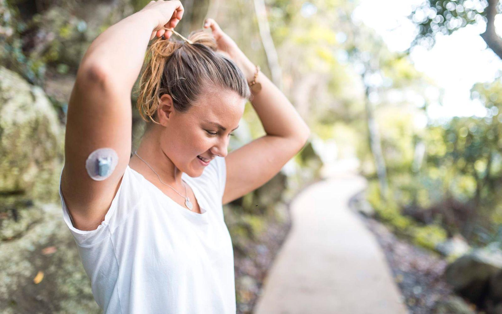 Diabetes: Frau mit Diabetessensor am Arm macht sich einen Zopf in der Natur.