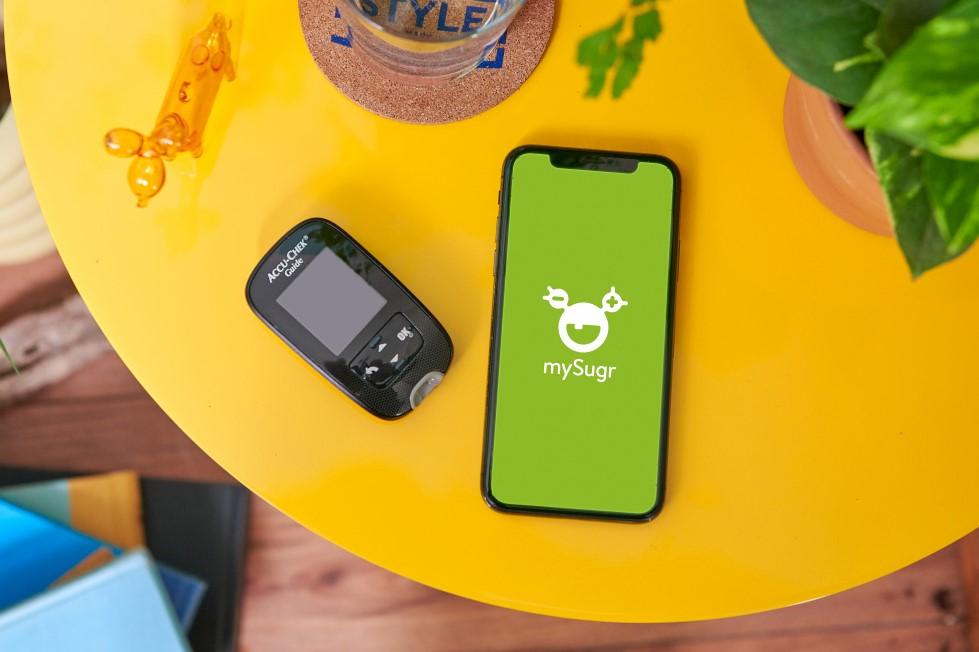Diabetes-App mySugr: Blutzuckermessgerät und Smartphone mit mySugr-App liegen auf einem runden gelben Tisch.