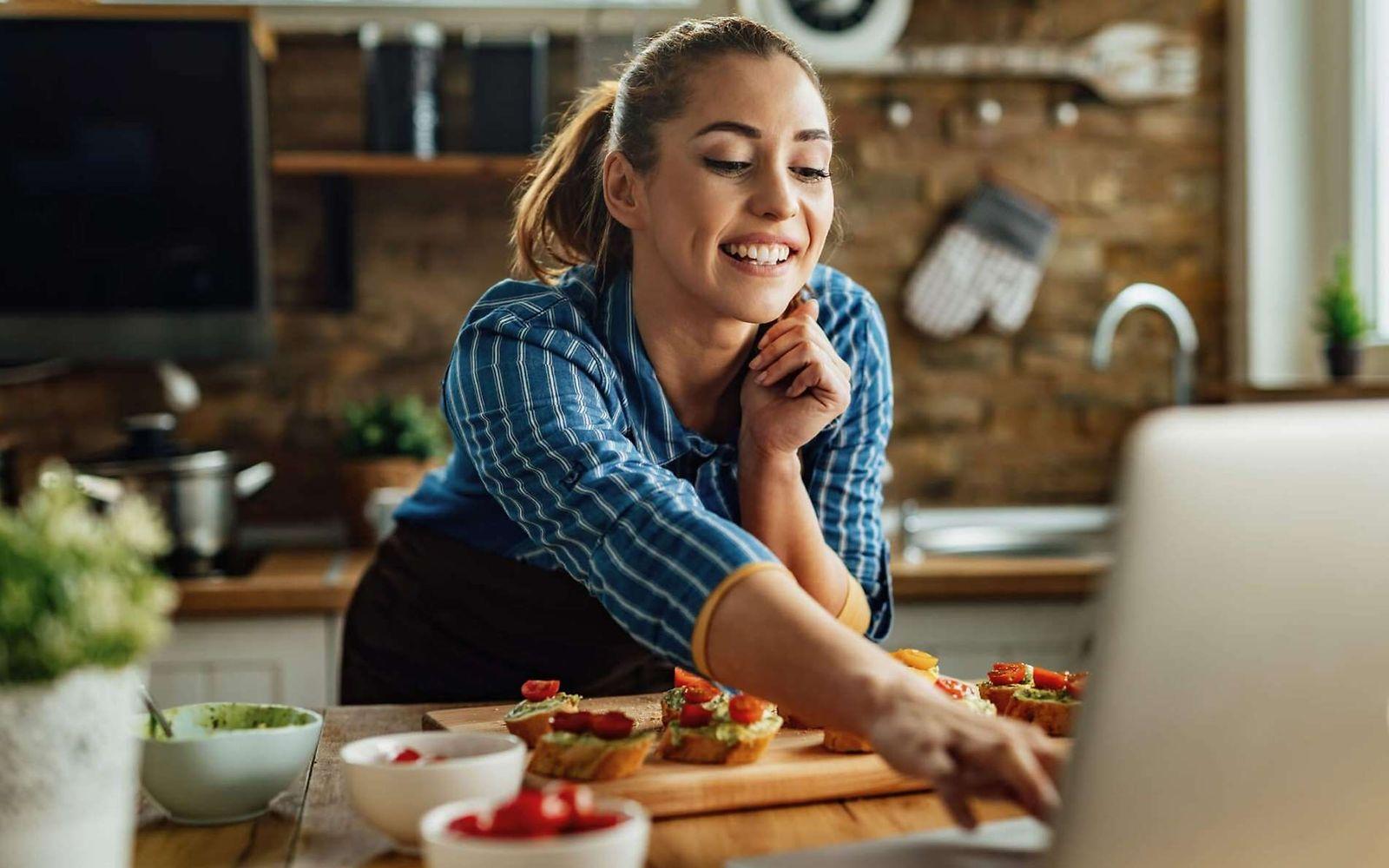 Natürlich ernähren: Junge Frau steht mit Essen und Laptop an der Küchenzeile