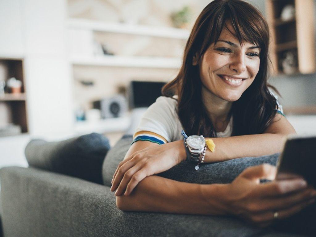 Frau mittleren Alters sitzt auf einem Sofa und schaut fröhlich auf Ihr Smartphone.