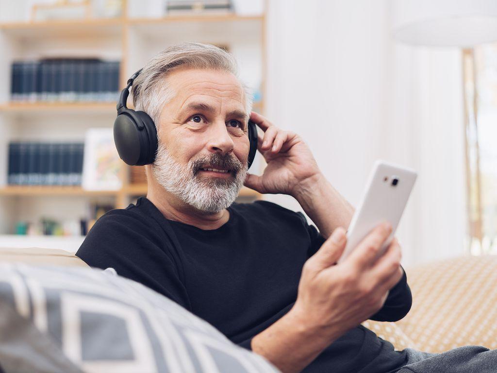 Älterer Mann sitzt auf dem Sofa, hat Kopfhörern auf und sein Handy in der Hand.