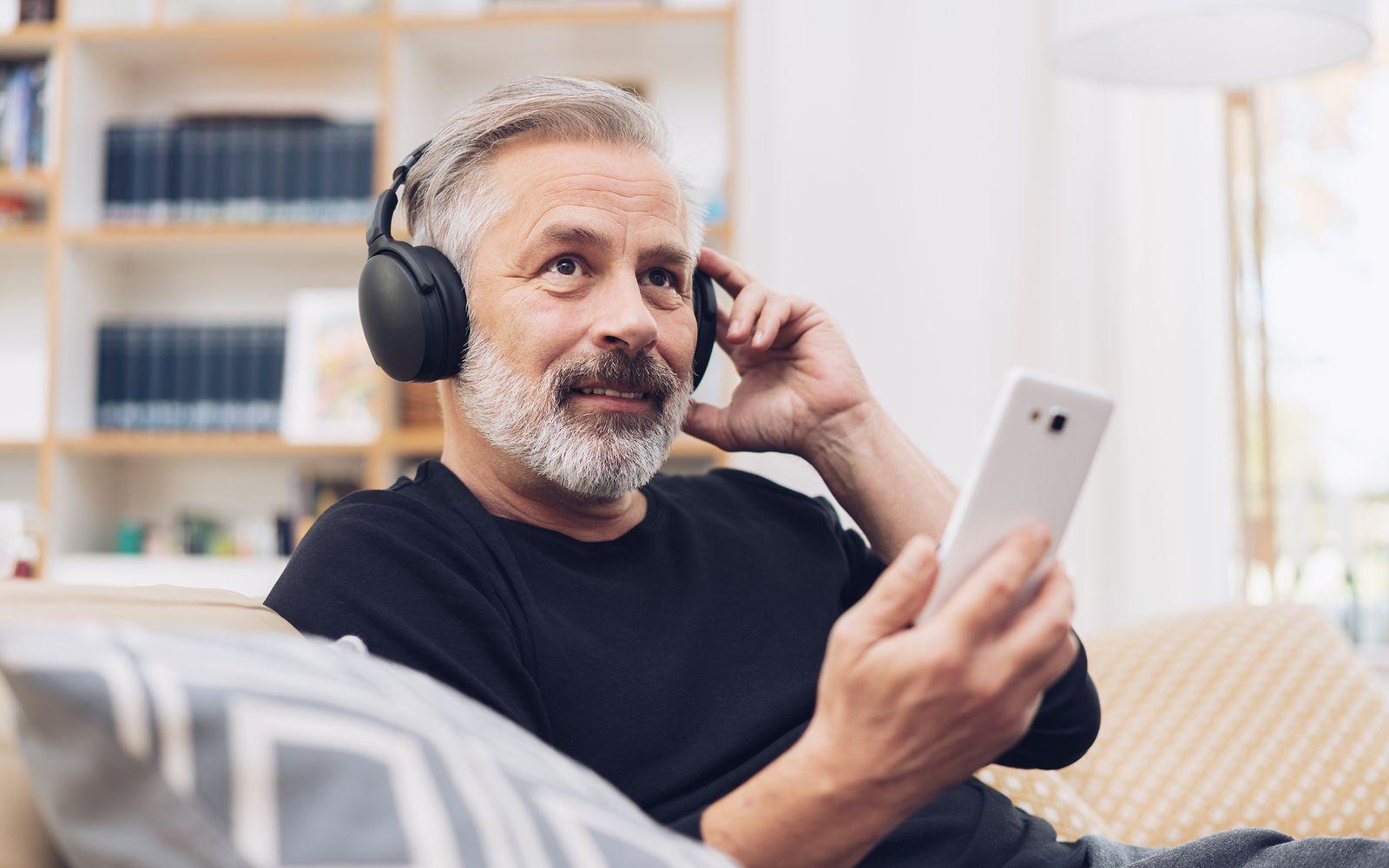 Älterer Mann sitzt auf dem Sofa, hat Kopfhörern auf und sein Handy in der Hand.