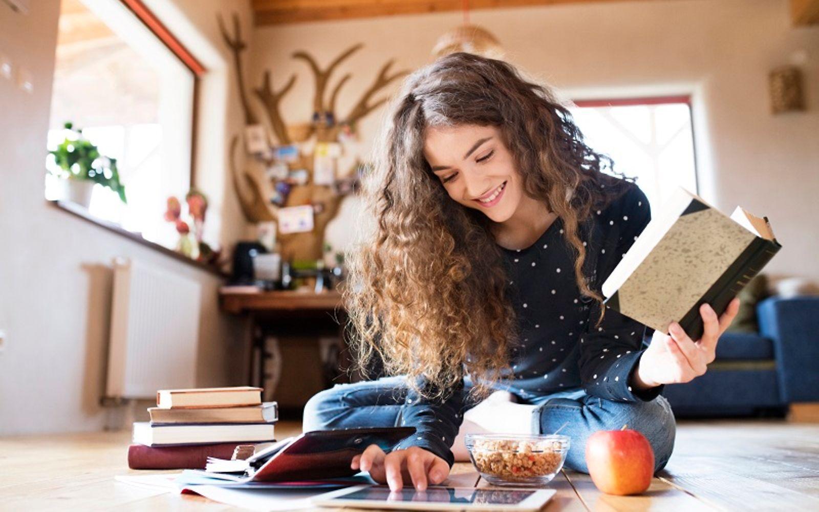 Bild: Jugendliche mit Buch in der Hand sitzt auf dem Boden und bedient ein Tablet.