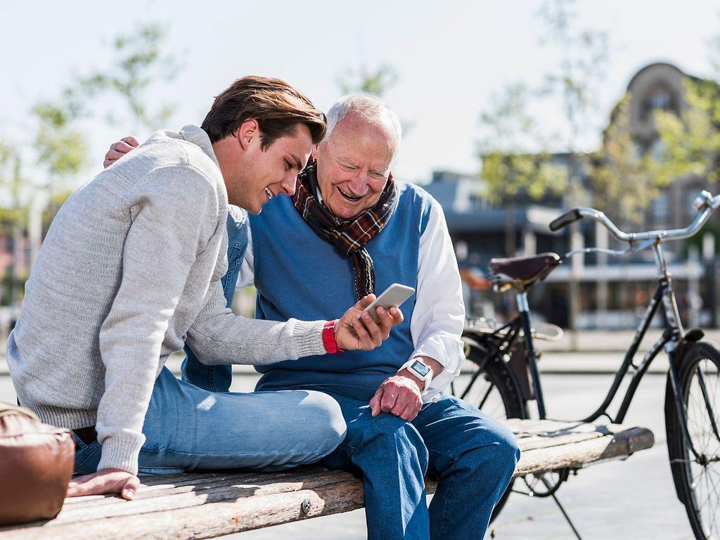 DAK Pflege-App: Enkel sitzt mit Großvater auf einer Parkbank und zeigt lachend auf das Smartphone