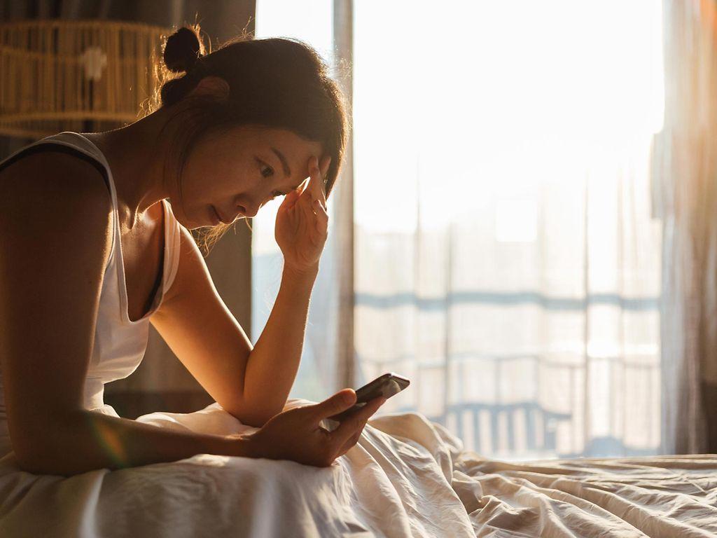 Fimo App bei Long Covid: Junge Frau sitzt erschöpft im Bett und schaut auf ihr Handy.