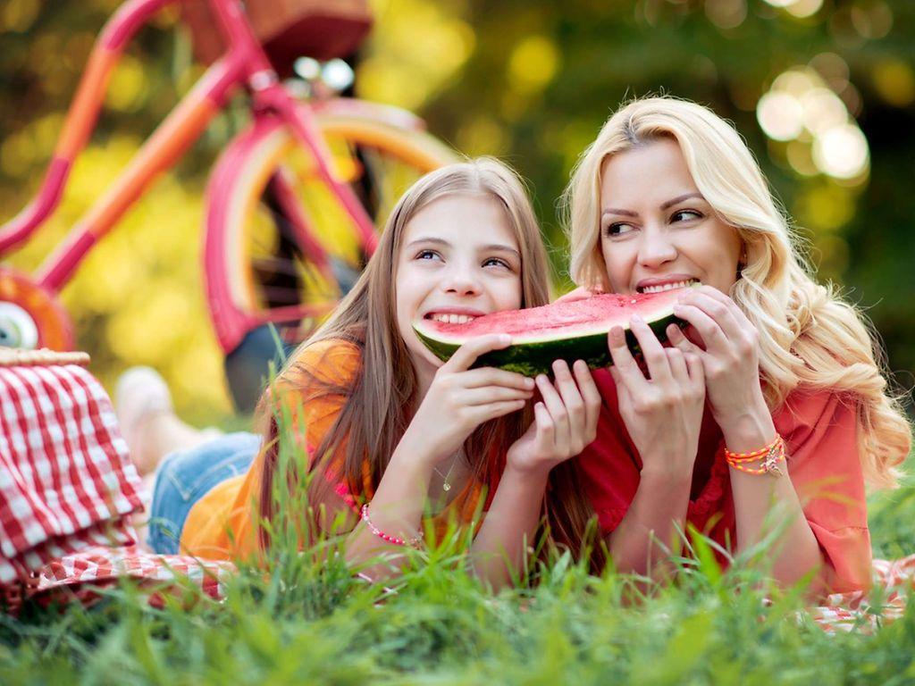 Ernährung bei Hitze: Mutter und Tochter liegen im Sommer im Park und essen eine Wassermelone.