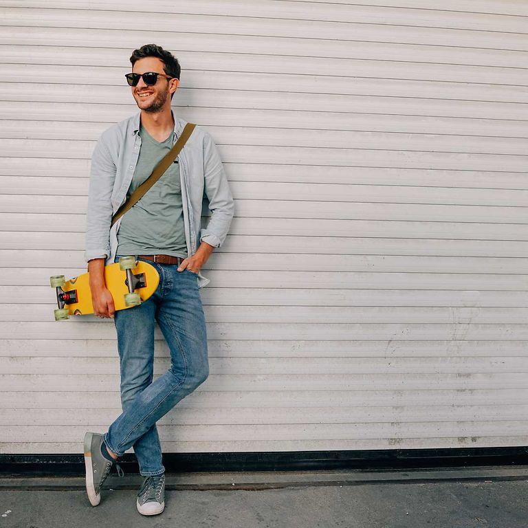 Bild: Junger Mann mit Sonnenbrille steht lässig an einer Wand und hat ein Skateboard unterm Arm.