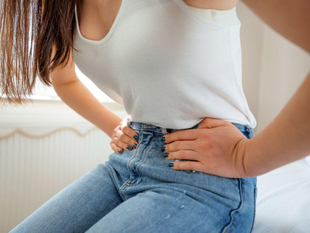 Symbolbild für Blasenentzündung: Eine Frau sitzt auf einem Bett und hält sich den Bauch