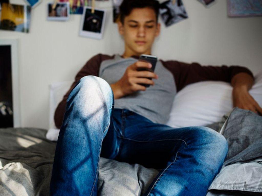 Symbolbild Pubertät bei Jungen: Ein Teenager sitzt auf einem Sofa und guckt auf ein Smartphone