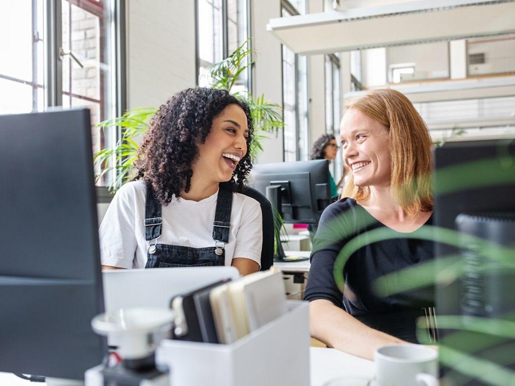 Bild: Zwei Frauen sitzen vor ihren PCs und lächeln sich an.