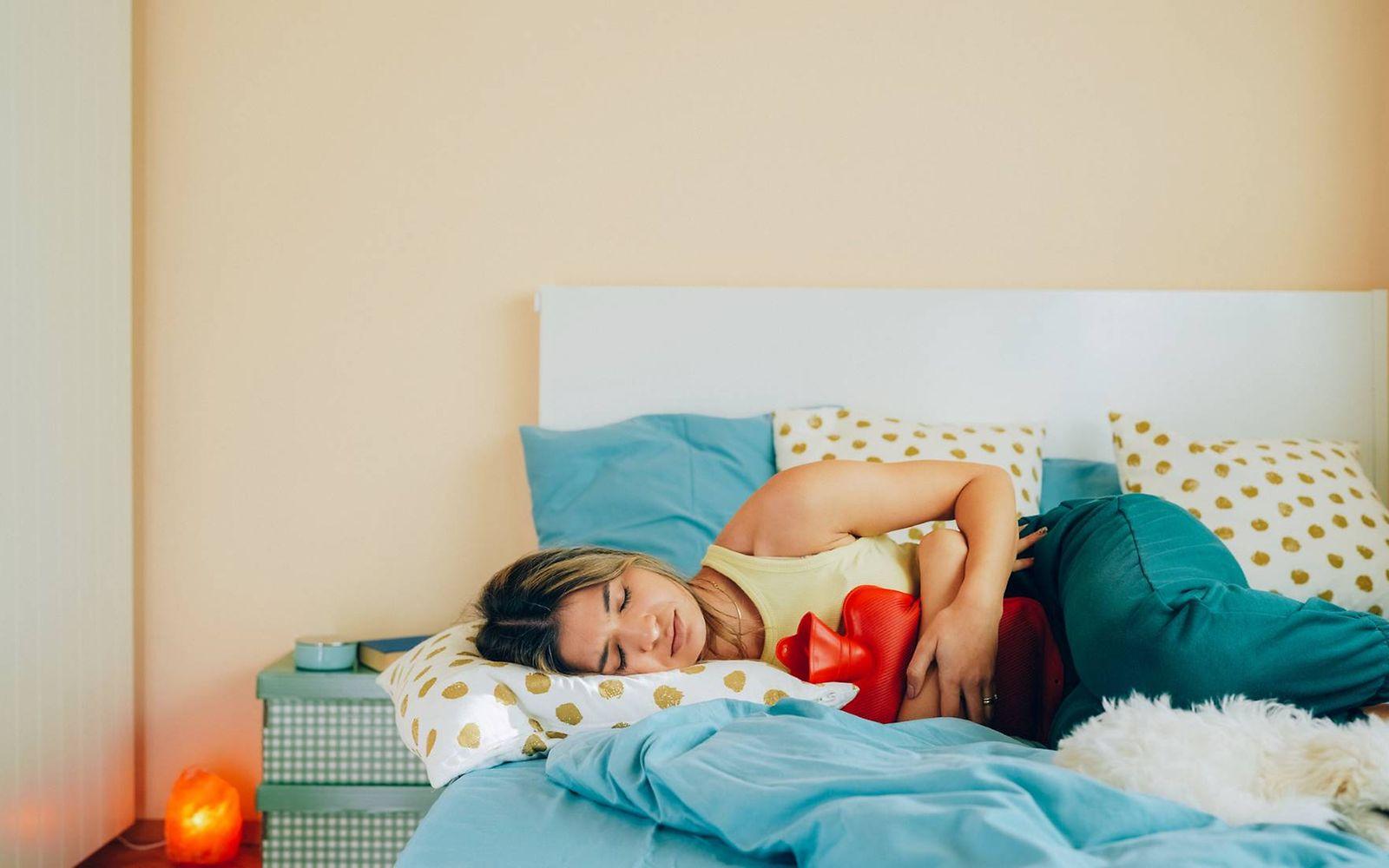 Unterleibsschmerzen: Junge Frau liegt mit Wärmflasche im Bett