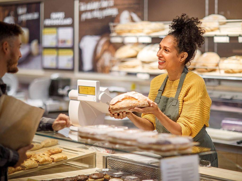 Krankenversicherung im Minijob: Freundliche Bäckereiverkäuferin präsentiert einem Kunden Brotlaib 