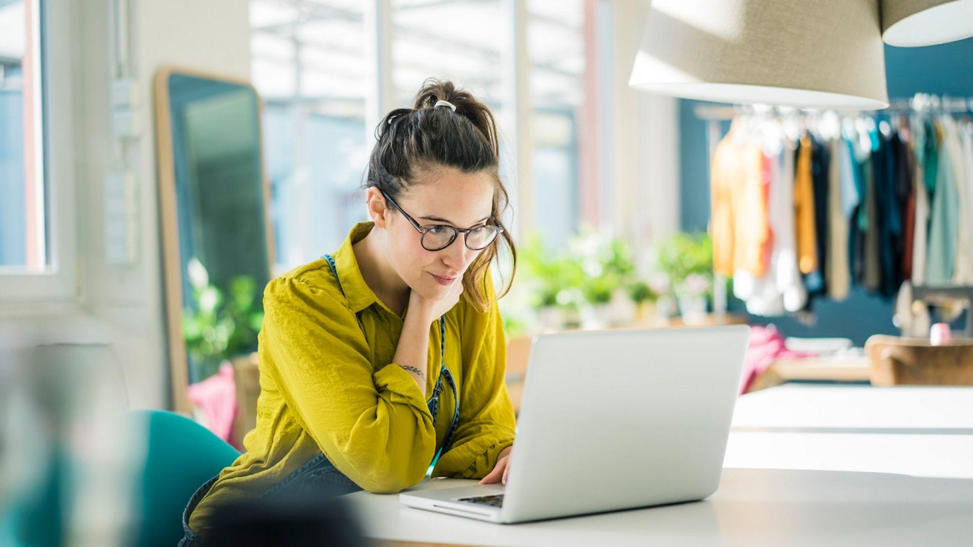 Junge Frau mit Brille sitzt am Schreibtisch und schaut in den Laptop.