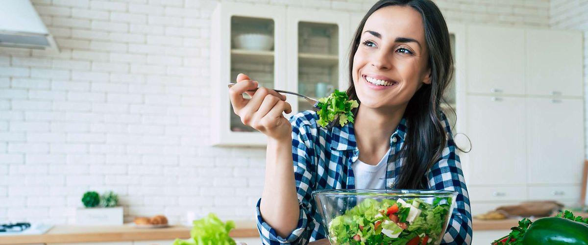 Fit & Cash: Junge Frau sitzt in ihrer Küche und isst einen Salat.