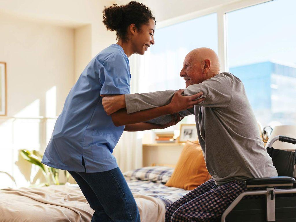 Bild: Pflegerin hilft einem älteren Mann zuhause aus seinem Rollstuhl