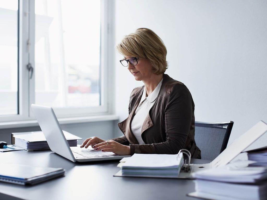 Bild: Frau mittleren Alters sitzt im Büro am Laptop und hat Akten neben sich.