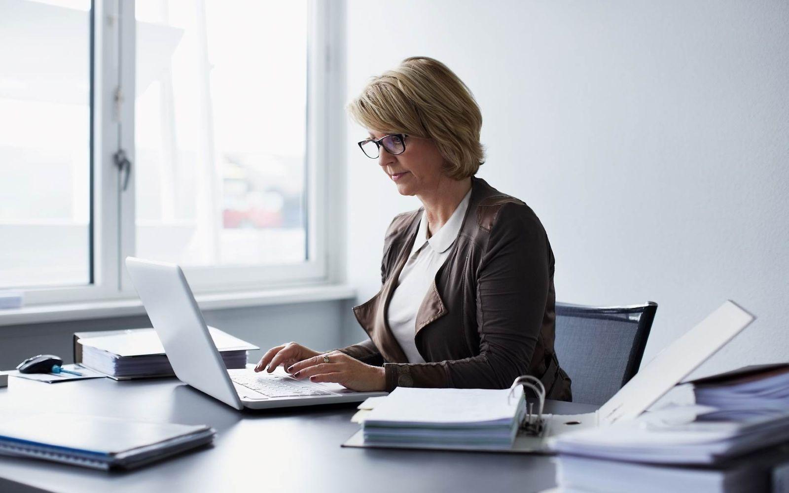 Bild: Frau mittleren Alters sitzt im Büro am Laptop und hat Akten neben sich.