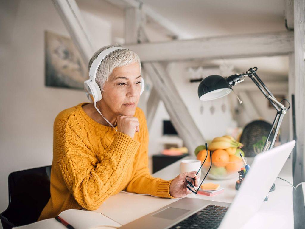 Bild: Frau mit gelben Pullover sitzt am Laptop und hört einer Veranstaltung zu.