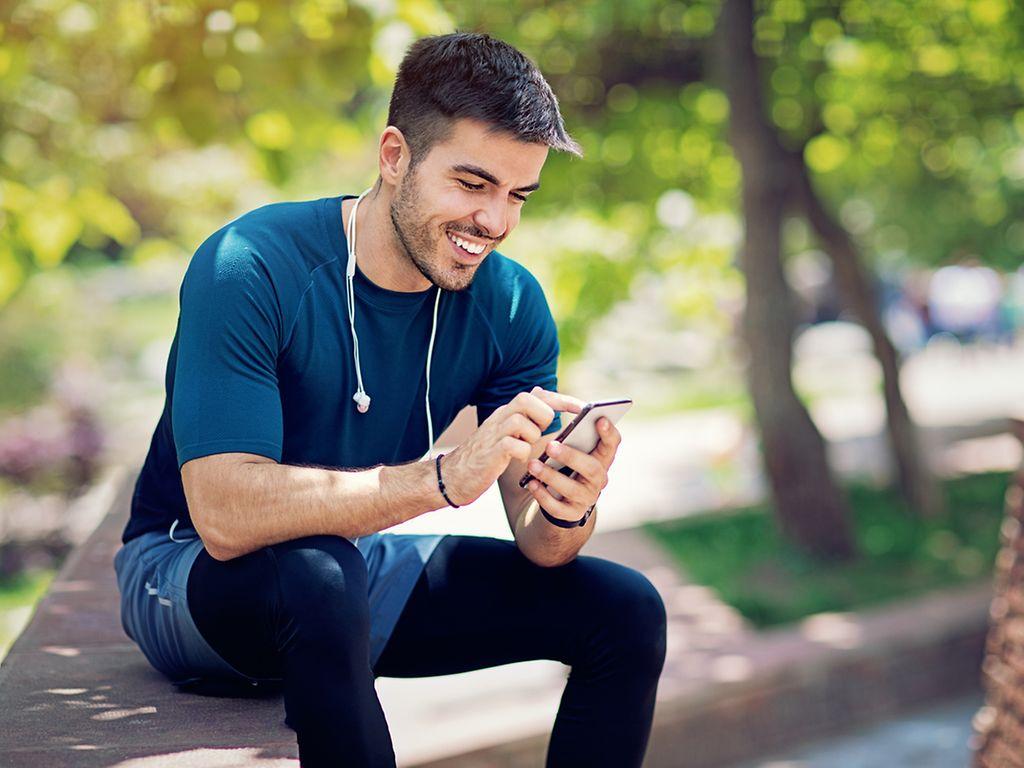 Symbolbild: Junger sportlich aussehender Mann blickt lächelnd auf seinSmartphone.