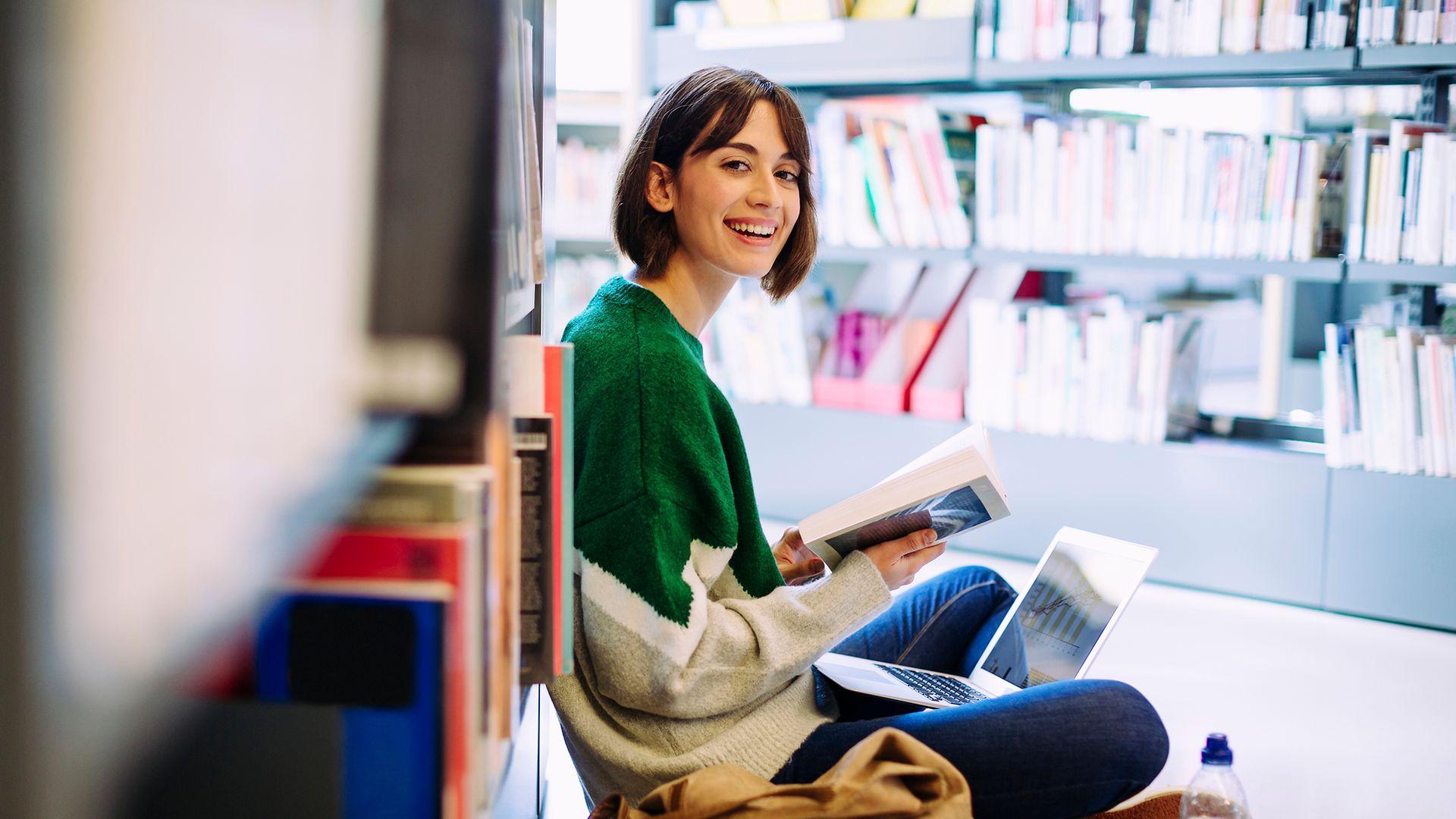 Portrait of woman with laptop computer reading book while sitting on floor in library.