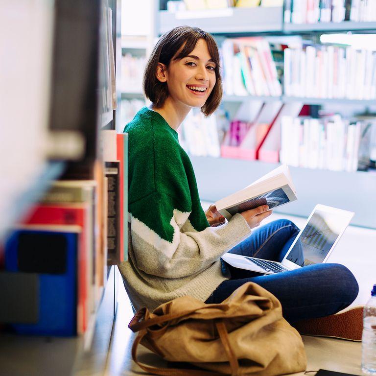 Portrait of woman with laptop computer reading book while sitting on floor in library.