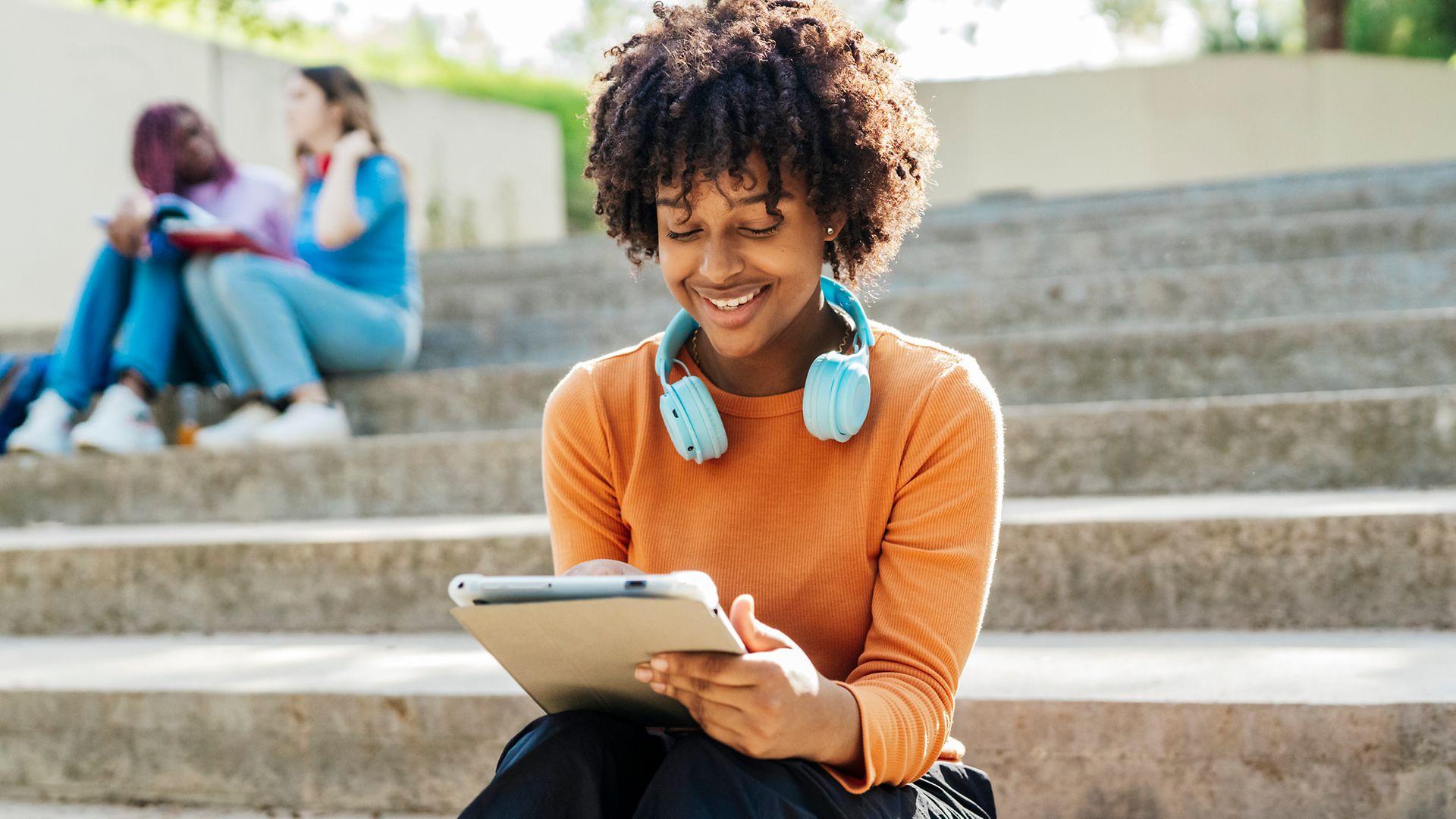 Young woman is sitting on stairs with blue headphones around her neck and looking at a digital tablet while smiling.