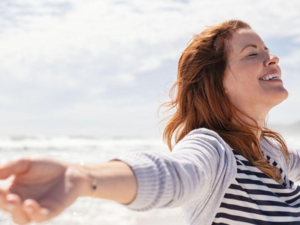 Kraft der Affirmationen: Frau steht lächelnd am Meer und genießt die Sonne und den Wind.