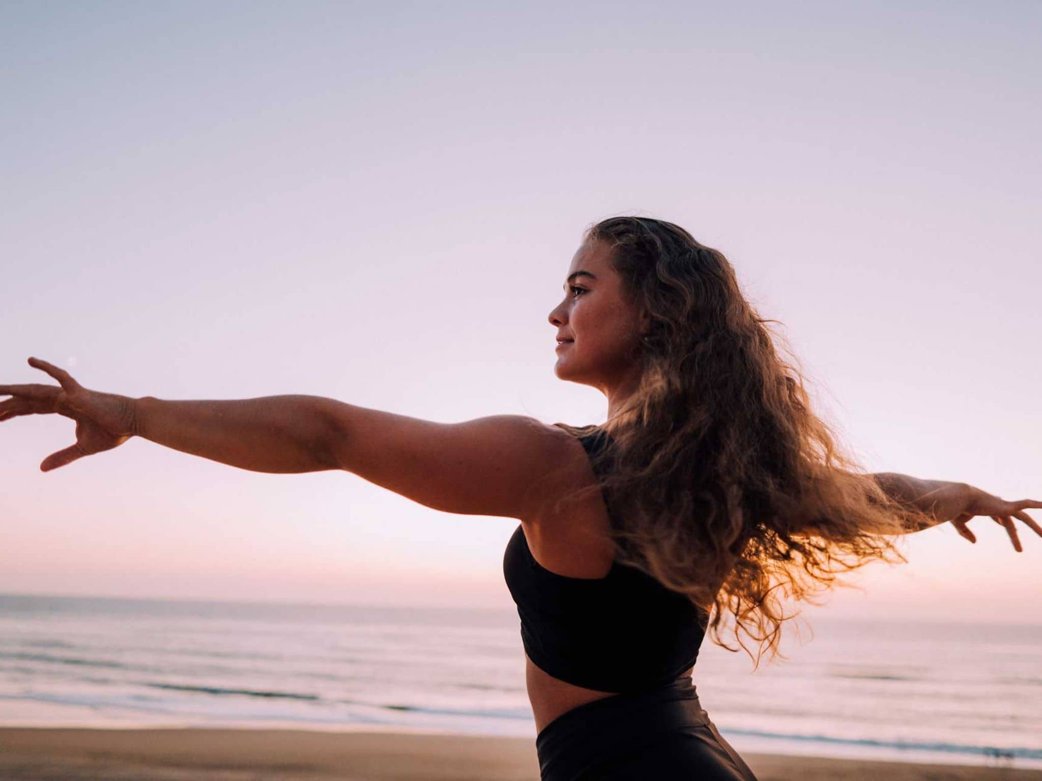 Junge Frau blickt fröhlich mit ausgebreiteten Armen über den Strand