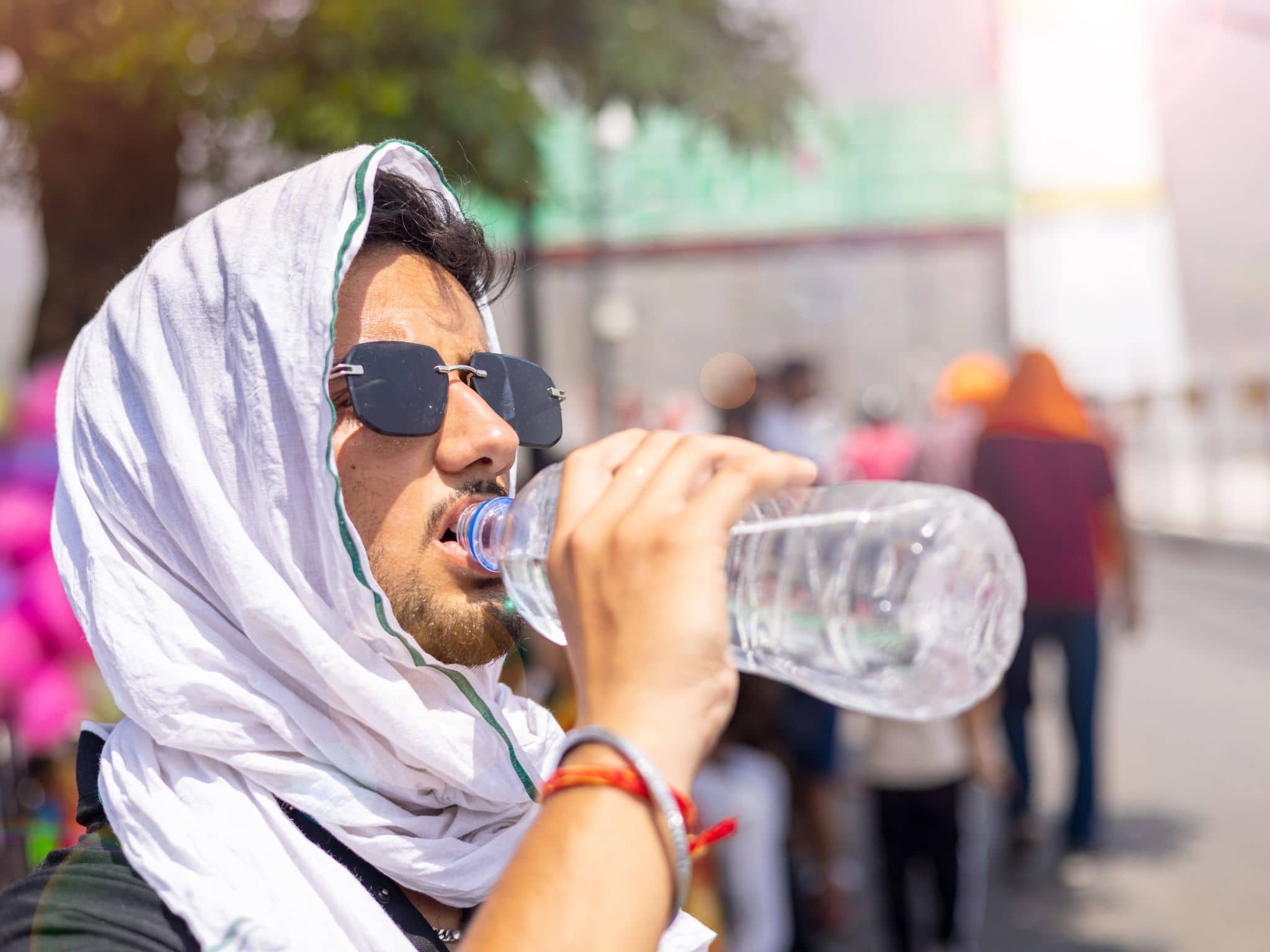 Junger Mann schützt sich vor Sonne und Hitze mit einem weißen Kopftuch und trinkt Wasser aus einer Flasche.