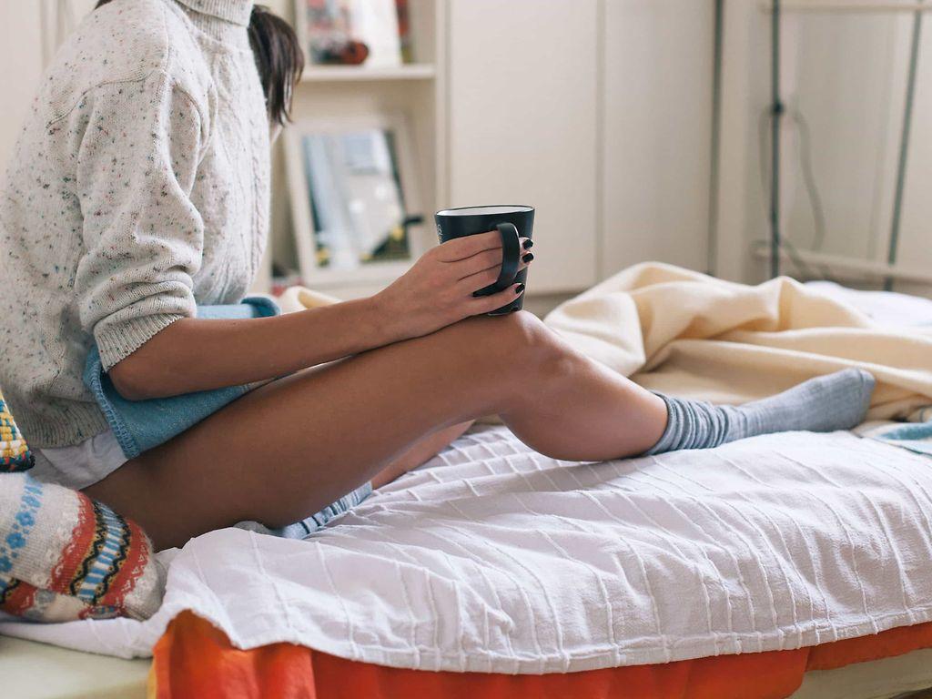 Menstruationsbeschwerden und PMS: Frau sitzt mit nackten Beinen, Wärmflasche und Teebecher auf einem Bett.