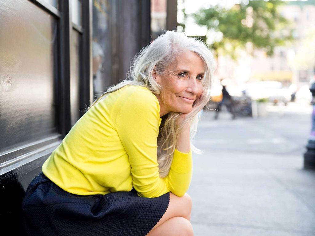 Wechseljahresbeschwerden: Ältere, draußen sitzende Frau stützt lächelnd den Kopf auf die Hand. 